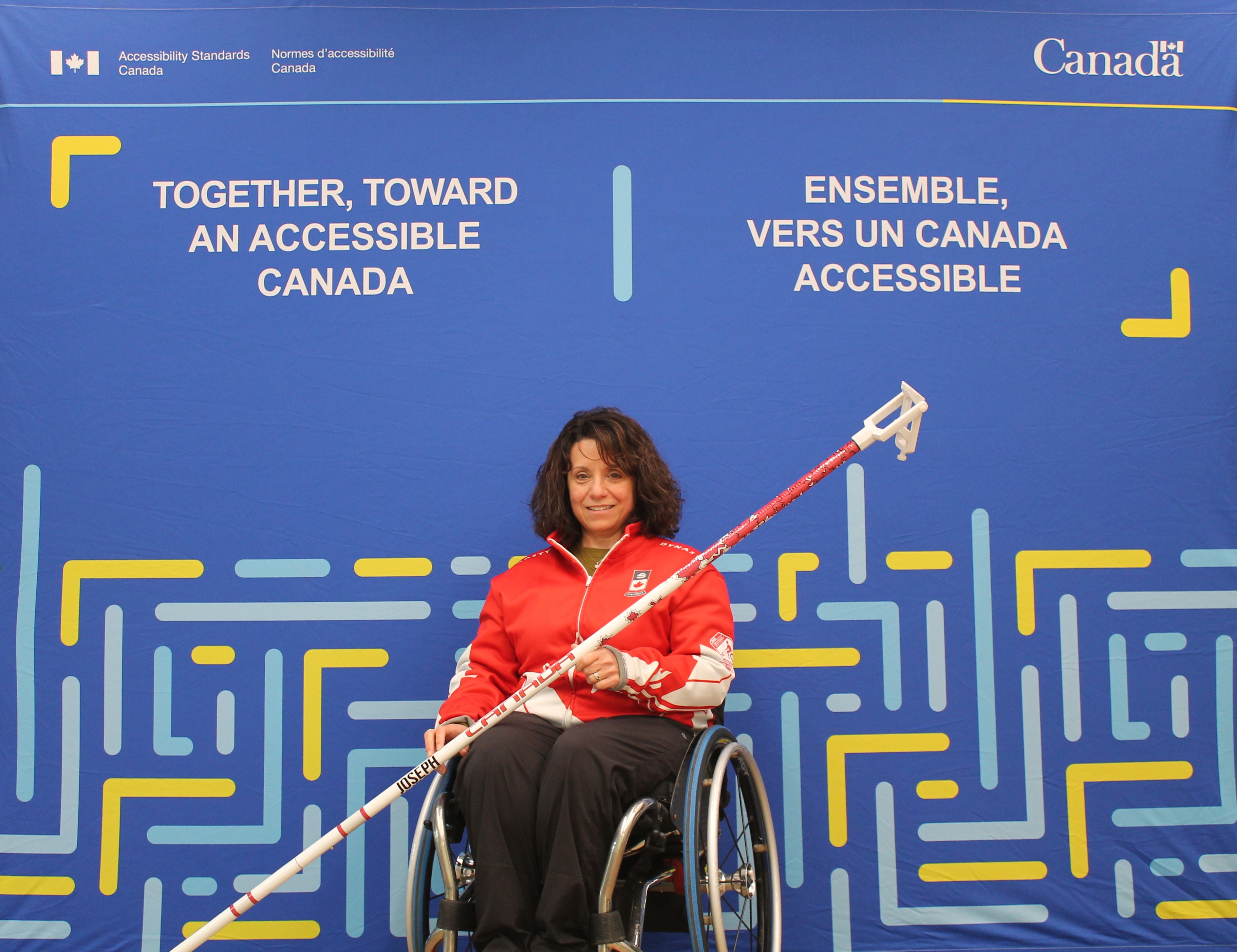 Collinda Joseph pose avec son fauteuil roulant et son bâton de curling. Le bâton est muni d'une poignée réglable à l'extrémité qui saisit la pierre et lui permet de la lancer à une certaine distance. Elle se trouve devant une toile de fond bleue sur laquelle est apposé le logo des Normes d'accessibilité Canada. Le slogan en anglais se lit comme suit : "Together, toward an accessible Canada". Le slogan français se lit comme suit : "Ensemble vers un Canada Accessible".