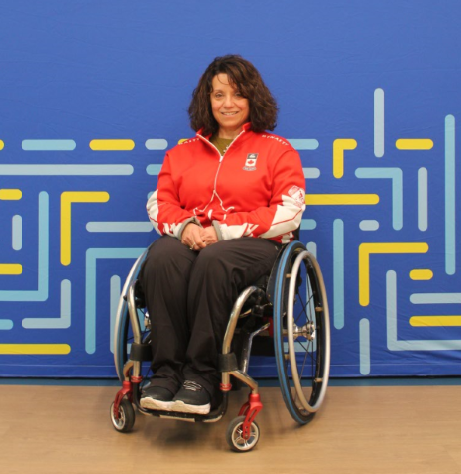 Portrait of Collinda Joseph. She is in her wheelchair, wearing her Team Canada jacket. She is in front of the blue and yellow Accessibility Standards Canada banner.