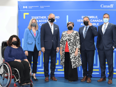 Six individus avec des masques de visage sont devant un fond bleu. Ils représentent deux organisations : Normes d'accessibilité Canada et l'International Code Council.