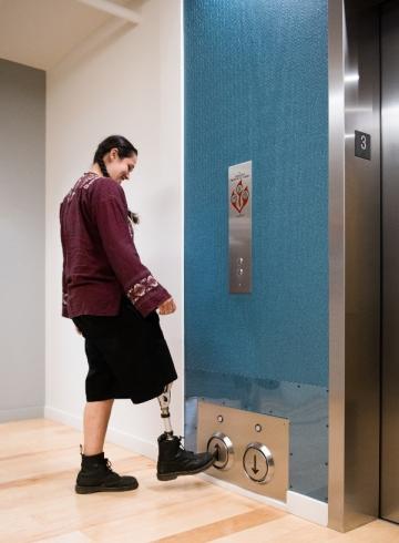 Une personne autochtone bispirituelle appelle l'ascenseur en appuyant sur le bouton de l'étage avec sa jambe prothétique.